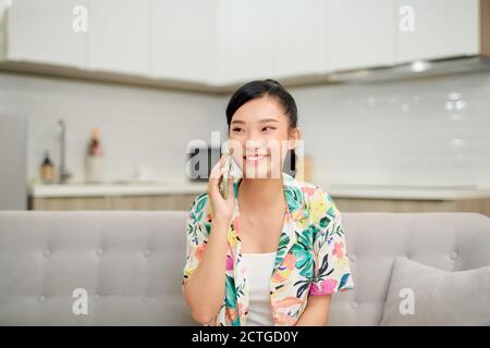 Angenehmes Gespräch mit Freund. Attraktive lächelnde junge Rothaarige Frau, die auf dem Handy spricht, während sie zu Hause auf dem beigefarbenen Sofa sitzt Stockfoto