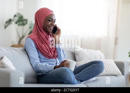 Entspannendes Wochenende. Fröhliche Schwarze Muslimische Frau Im Gespräch Auf Handy Und Genießen Kaffee Stockfoto