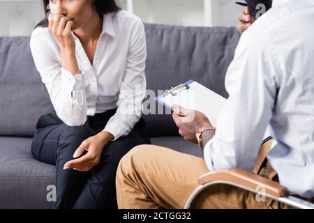 Beschnittene Ansicht einer besorgten Frau, die die Hand in der Nähe des Gesichts hält Sitzen in der Nähe afroamerikanischen Psychologen hält Zwischenablage Stockfoto