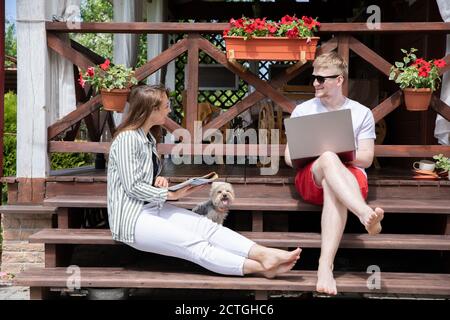 Junge, moderne Paare, die fern von zu Hause arbeiten, sitzen auf der Veranda Des Landhauses Stockfoto