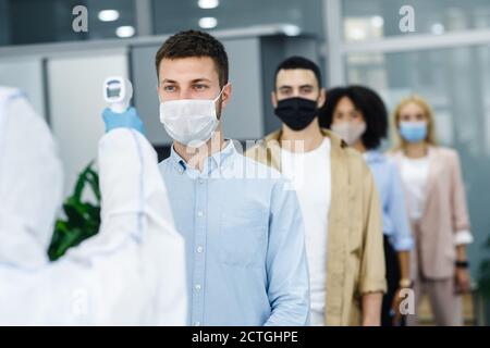 Morgen bei der Arbeit während covid-19 Epidemie. Person mit berührungslosem Thermometer misst die Temperatur von multirassischen Mitarbeitern in Schutzmasken Stockfoto