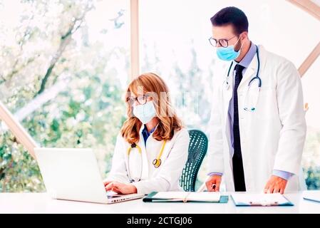 Aufnahme des medizinischen Teams, das in der Arztpraxis zusammenarbeitet. Ärztin sitzt hinter ihrem Laptop, während der Arzt neben ihr steht und diskutiert Stockfoto