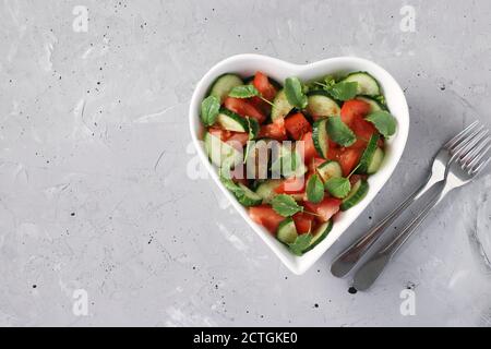 Herzförmige Schüssel mit gesundem Salat mit Tomaten, Gurken, Rucola und Rettich-Microgreens auf grauem Beton-Hintergrund, gesunde Ernährung Tag Stockfoto