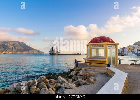Pigadia, Karpathos, Griechenland - 4. Oktober 2019: Panorama am frühen Morgen Blick auf verlassene Tourist Info Center mit roter Kuppel und angedocktem Frachtschiff Stockfoto