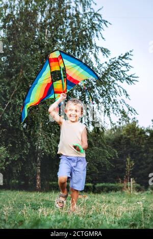 Fröhlicher kleiner Junge läuft mit einem bunten Drachen auf der grünen Wiese des Stadtparks. Lustige Kindheit Konzept Bild. Stockfoto