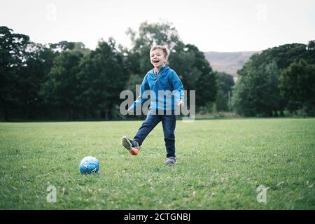 Ein vierjähriger Junge lacht, als er in einem Park in Ilkley, West Yorkshire, Großbritannien, einen Ball schießt. Stockfoto