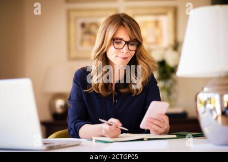 Lächelnde, reife Geschäftsfrau, die am Schreibtisch sitzt und SMS-Nachrichten mitgibt, während sie von zu Hause aus arbeitet. Heimbüro. Stockfoto