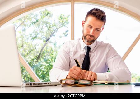 Ein gutaussehender Geschäftsmann, der am Schreibtisch am Fenster sitzt und ein paar Papiere macht. Stockfoto