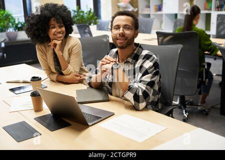 Junger fröhlicher gemischter Renner mit Brille, Blick in die Kamera und lächelnd, sitzt mit seiner fröhlichen Kollegin im modernen Coworking Space am Schreibtisch. Geschäftsleute arbeiten zusammen Stockfoto