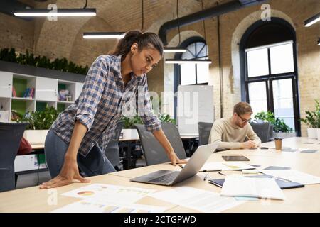 Fokussierte Mixed Race junge Geschäftsfrau, die im modernen Coworking Space steht, auf den Laptop-Bildschirm schaut und über etwas nachdenkt, während ihr männlicher Mitarbeiter im Hintergrund arbeitet. Geschäftsleute Stockfoto
