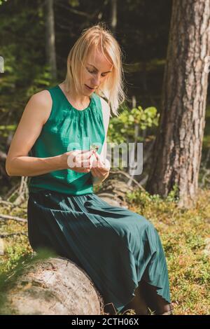 Blonde Frau mit grüner Bluse und Kleid mit geschlossenen Augen Auf altem Baumstamm sitzend und in den Händen halten Cranberry Zweig vor dicht und dunkel fo Stockfoto