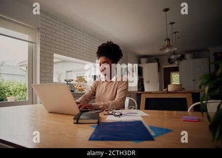 Junge konzentrierte afrikanische Frau mit lockigen Haaren arbeitet von zu Hause aus Am Computer, während Sie zu Hause am Tisch sitzen Stockfoto