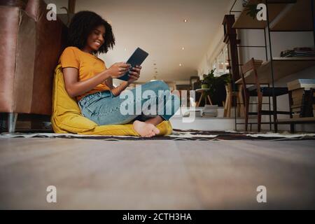 Low-Winkel-Ansicht der glücklichen afroamerikanischen Frau mit Afro Sitzen auf dem Boden mit einem Kissen mit einem digitalen Tablet Stockfoto