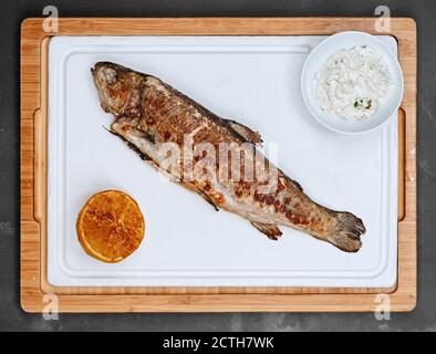 Gesund geröstete Portion von Seebarsch ganzen Fisch mit der Hälfte Zitrone und Sauce auf weißem Tablett und Holzplatte Anzeigen Stockfoto