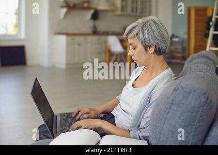 Seriöse ältere Frau, die am Laptop arbeitet, sitzt in einem großen Sessel Mit Notebook daneben Stockfoto