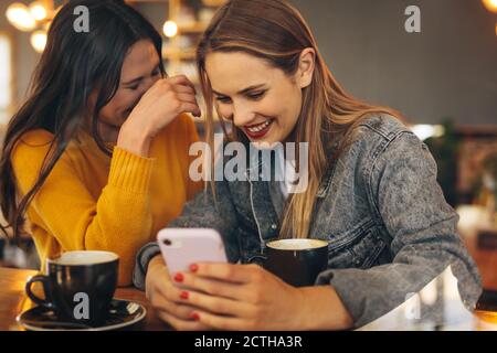 Frau, die ihrem Freund etwas Lustiges auf ihrem Handy zeigt und lächelt. Zwei Freundinnen sitzen an einem Tisch im Café und schauen auf ein Handy und