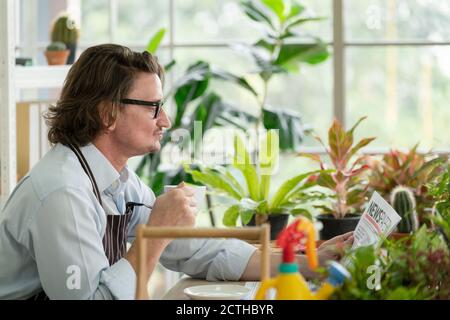 Mann in legerem Outfit mit Brille sitzend und Kaffee trinkend Frühstück beim Lesen der Zeitung im modernen grünen Gartenhaus Stockfoto