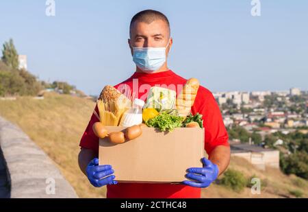 Lebensmittelzustellung, Spende oder Unterstützung während der Quarantäne. Männlicher Kurier in rotem T-Shirt mit Karton mit Lebensmitteln Stockfoto