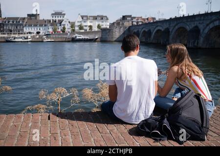 An einem Sommertag sitzt ein junges Paar auf einer Mauer an der Maas in Maastricht mit der mittelalterlichen St. Servatius Brücke im Hintergrund Stockfoto
