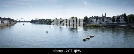 Kinder spielen mit Booten auf der Maas, im Hintergrund die Fußgängerbrücke Hoge Brug und rechts der Kai mit alten Gebäuden in Maastri Stockfoto