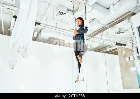 Ariel Yoga. Junge kaukasische Frau in Sportkleidung, die im Studio Fliegenyoga praktiziert, auf dem einen Bein in der weißen Hängematte stehend und meditierend, volle Länge. Wellness und gesunder Lebensstil Stockfoto