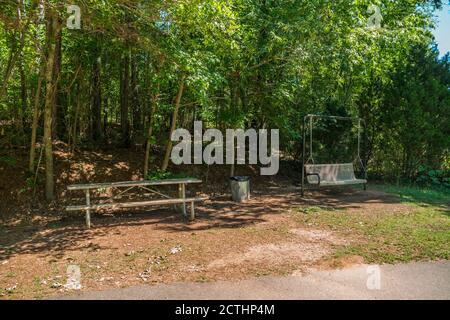 Leerer Picknicktisch und eine Sitzschaukel mit einem Metall Mülleimer unter den Bäumen für Schatten neben dem Wandern Wanderwege in einem bewaldeten Park an einem sonnigen Tag Stockfoto