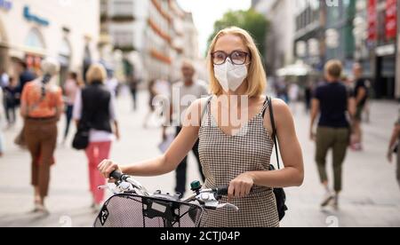 Frau, die mit ihrem Fahrrad auf der Fußgängerstraße in der Stadt unterwegs ist und in der Öffentlichkeit eine medizinische Gesichtsmaske trägt, um die Verbreitung des Corona-Virus zu verhindern. Neuer Normalwert während Stockfoto