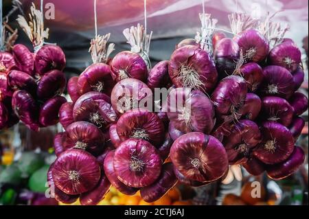 Gesammelte rote Zwiebeln in Zöpfen hängen am Marktfenster. Die Handernte wird auf dem Bauernmarkt verkauft Stockfoto