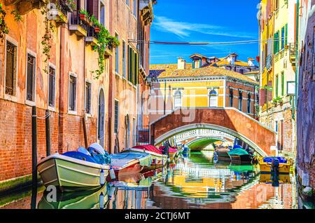 Venedig Stadtbild mit Gondeln und Motorbooten, die auf einem schmalen Wasserkanal in der Nähe von bunten Gebäuden und Steinbrücke festgemacht sind, Region Venetien, Norditalien, blauer Himmel am Sommertag, typisch venezianischer Blick Stockfoto