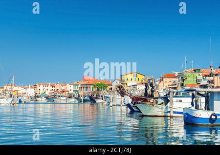 Fischerschiffe und Boote in der Hafenlagune in der Nähe der Küste von Sottomarina Stadt mit Reihe von bunten Gebäuden im Sommer Tag, blauer Himmel Hintergrund, Region Venetien, Norditalien Stockfoto