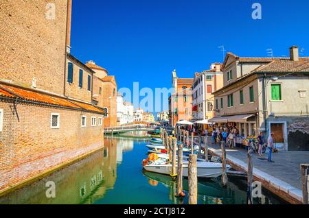 Chioggia, Italien, 16. September 2019: Schmaler Wasserkanal Vena mit bunten Booten zwischen alten bunten Gebäuden und Backsteinbrücke, blauer Himmel Hintergrund im Sommer, Region Venetien Stockfoto