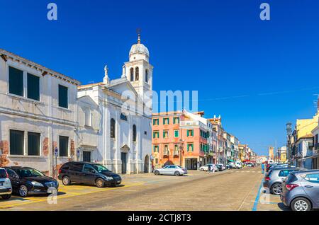 Chioggia, Italien, 16. September 2019: Katholische Kirche Chiesa di San Francesco, Reihe von bunten bunten bunten Gebäuden, Parkplatz Autos auf der Hauptstraße in der Altstadt, blauer Himmel im Sommer Tag Stockfoto