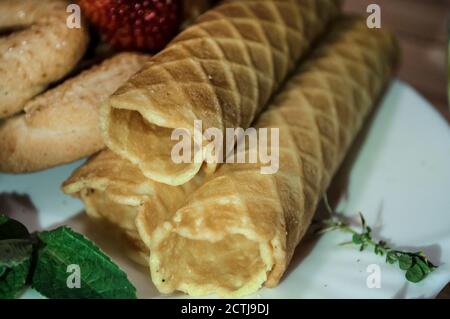 Waffelbrötchen und Kekse auf einem weißen Teller und eine Tasse Tee. Waffeln auf der Seite. Komposition auf einem hölzernen Hintergrund. Sichtbare Waffelstruktur Stockfoto