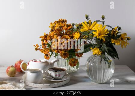 Ein Bouquet von orangefarbenem helenium mit wilden Traubenblättern in einer geriffelten Glasvase an einer dunkelblauen Wand. Weiße Teekanne, Tasse und Untertasse und Milchkännchen für Tee Stockfoto