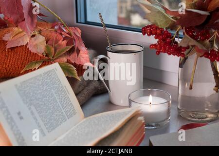 Eine eiserne Tasse Kaffee mit einem Löffel auf der Fensterbank zwischen einer Vase mit Zweigen und Viburnum-Beeren, einem Pullover, einem offenen Buch und einer Kerze. Foto für Stockfoto