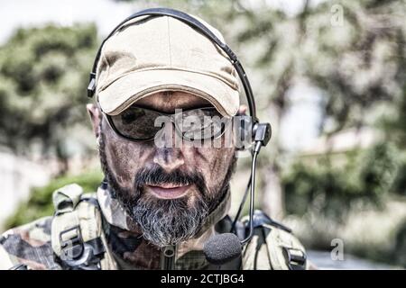 Schulterportrait des Soldaten, Spezialeinsatzkämpfers, moderner Kampfkämpfer mit schmutzigem, unrasierten Gesicht, Sonnenbrillen, Kampfhelm und Sprechern im taktischen Radio-Headset während der Mission Stockfoto