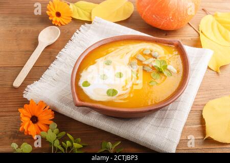 Traditionelle Kürbiscremesuppe mit Samen in Tonschüssel auf braunem Holzhintergrund mit Leinenserviette. Seitenansicht, Nahaufnahme. Stockfoto