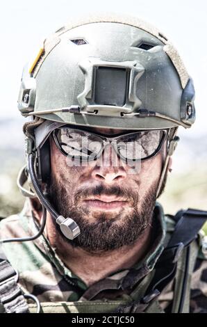 Schulterportrait des Soldaten, Spezialeinsatzkämpfers, moderner Kampfkämpfer mit schmutzigem, unrasierten Gesicht, Sonnenbrillen, Kampfhelm und Sprechern im taktischen Radio-Headset während der Mission Stockfoto