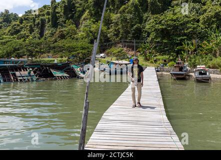 Ein junger Mann mit Kamera steht auf dem Holzsteg an den Stelzenhäusern von Pukat Village, einem Fischerdorf entlang Jalan Bokara in Sandakan, Sabah Stockfoto