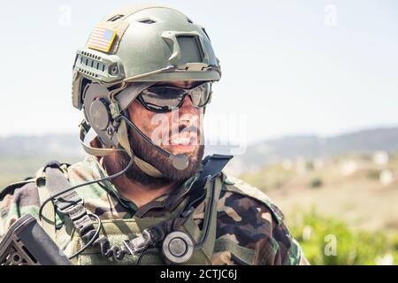 Schulterportrait des Soldaten, Spezialeinsatzkämpfers, moderner Kampfkämpfer mit schmutzigem, unrasierten Gesicht, Sonnenbrillen, Kampfhelm und Sprechern im taktischen Radio-Headset während der Mission Stockfoto