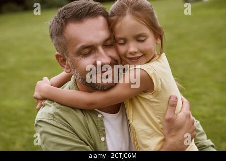 Portrait von jungen liebevollen Vater und sein nettes kleines Mädchen halten die Augen geschlossen und umarmt, während die Zeit verbringen outddors, sitzen auf einem grünen Gras Stockfoto