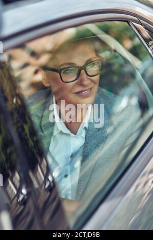 Vertikale Aufnahme eines schönen und glücklichen Unternehmens mittleren Alters Frau trägt Brillen Blick aus einem Autofenster und Lächelnd auf dem Rücken sitzend Stockfoto