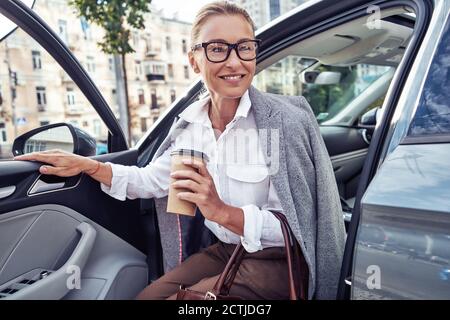Start Tag mit Kaffee. Schöne Geschäftsfrau mittleren Alters kam bei der Arbeit an, sie kommt aus ihrem modernen Auto und lächelt Stockfoto