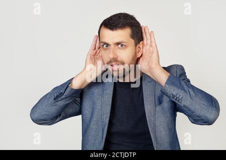 Junger Geschäftsmann Mann mit Bart versucht, beide Hände auf seinem Ohr zu hören, neugierig auf Klatsch. Hörprobleme, taub. Porträt eines Mannes auf grauem Hintergrund. Stockfoto
