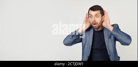 Junger Geschäftsmann Mann mit Bart versucht, beide Hände auf seinem Ohr zu hören, neugierig auf Klatsch. Hörprobleme, taub. Porträt eines Mannes auf grauem Hintergrund. Stockfoto