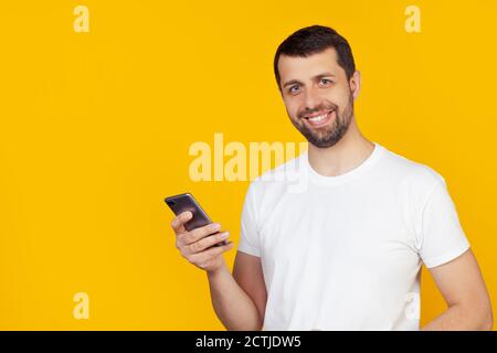 Der junge Mann mit einem Bart in einem weißen T-Shirt benutzt ein Smartphone mit einem glücklichen Gesicht, das steht und lächelt und ein selbstbewusstes Lächeln zeigt, das Zähne zeigt. Steht auf isoliertem gelben Hintergrund. Stockfoto