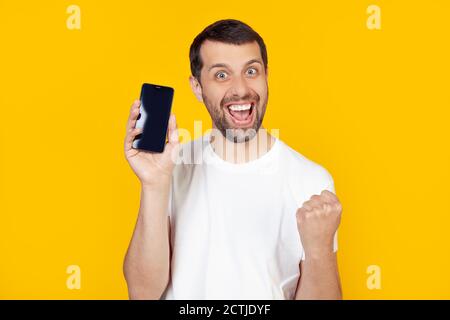 Ein junger Mann mit Bart in einem weißen T-Shirt zeigt einen Smartphone-Bildschirm, schreit mit Stolz und feiert Sieg und Erfolg, ist sehr aufgeregt, freut sich über Emotionen. Steht auf isoliertem gelben Hintergrund. Stockfoto