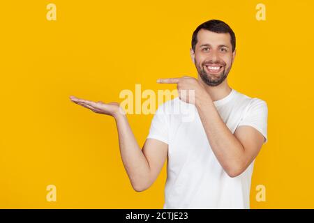 Der junge Mann mit Bart in einem weißen T-Shirt ist erstaunt und lächelt auf die Kamera, zeigt mit der Hand und zeigt mit dem Finger. Steht auf isoliertem gelben Hintergrund. Stockfoto