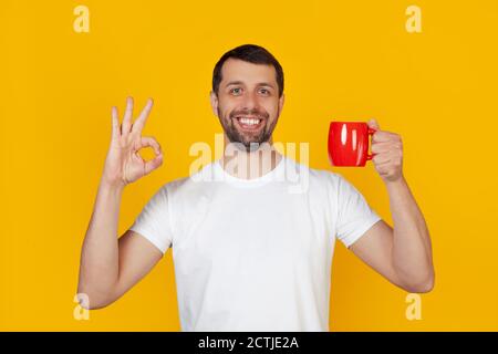 Junger Mann mit einem Bart in einem weißen T-Shirt lächelnd, hält eine rote Tasse Kaffee und zeigt die Geste, alles ist in Ordnung, ein Mann mit einem glücklichen Gesicht. Steht auf isoliertem gelben Hintergrund. Stockfoto