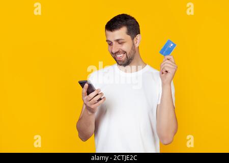 Junger Mann mit einem Bart in einem weißen T-Shirt mit einer Kreditkarte. Per Kreditkarte online bezahlen mit einem Smartphone. Steht auf isoliertem gelben Hintergrund. Stockfoto